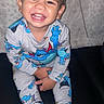 child, toddler, smiling, happy, portrait, sitting, pajamas, cartoon, colorful_clothing, bare_feet, couch, indoor, home, face, teeth, dark_hair, flash_photography, close_up, family, playful