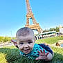 baby, blue_sky, casual_clothing, child, daytime, eiffel_tower, grass, greenery, happy, infant, landmark, nature, outdoor, park, person, reaching_hand, smiling, tourist_spot, travel, vacation