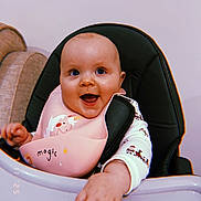 Sophia is registered to the contest to win money with this photo: baby, smiling, high_chair, bib, unicorn, magic, child, happy, infant, cute, indoor, person, clothing, reaching, hand, face, seat, joyful, expression, cozy