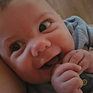 Ruben is registered to the contest to win money with this photo: baby, close_up, smiling, happy, infant, fingers, chewing, blue_clothing, face, head, skin, eyes, hand, cute, expression, indoors, child, person, portrait, wood_floor