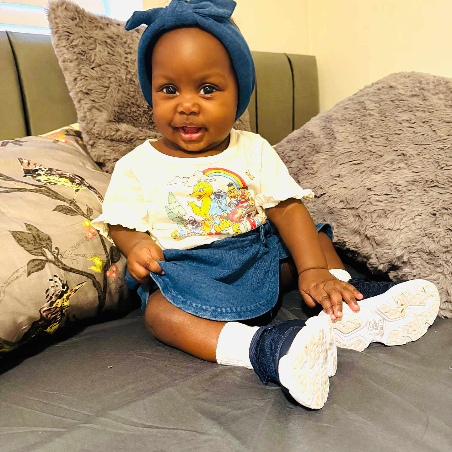 Dinorah is registered to the contest to win money with this photo: baby, bed, child, clothing, colorful, cozy, cute, footwear, furniture, happy, head_wrap, indoor, infant, person, pillow, portrait, shoes, sitting, smiling, tshirt