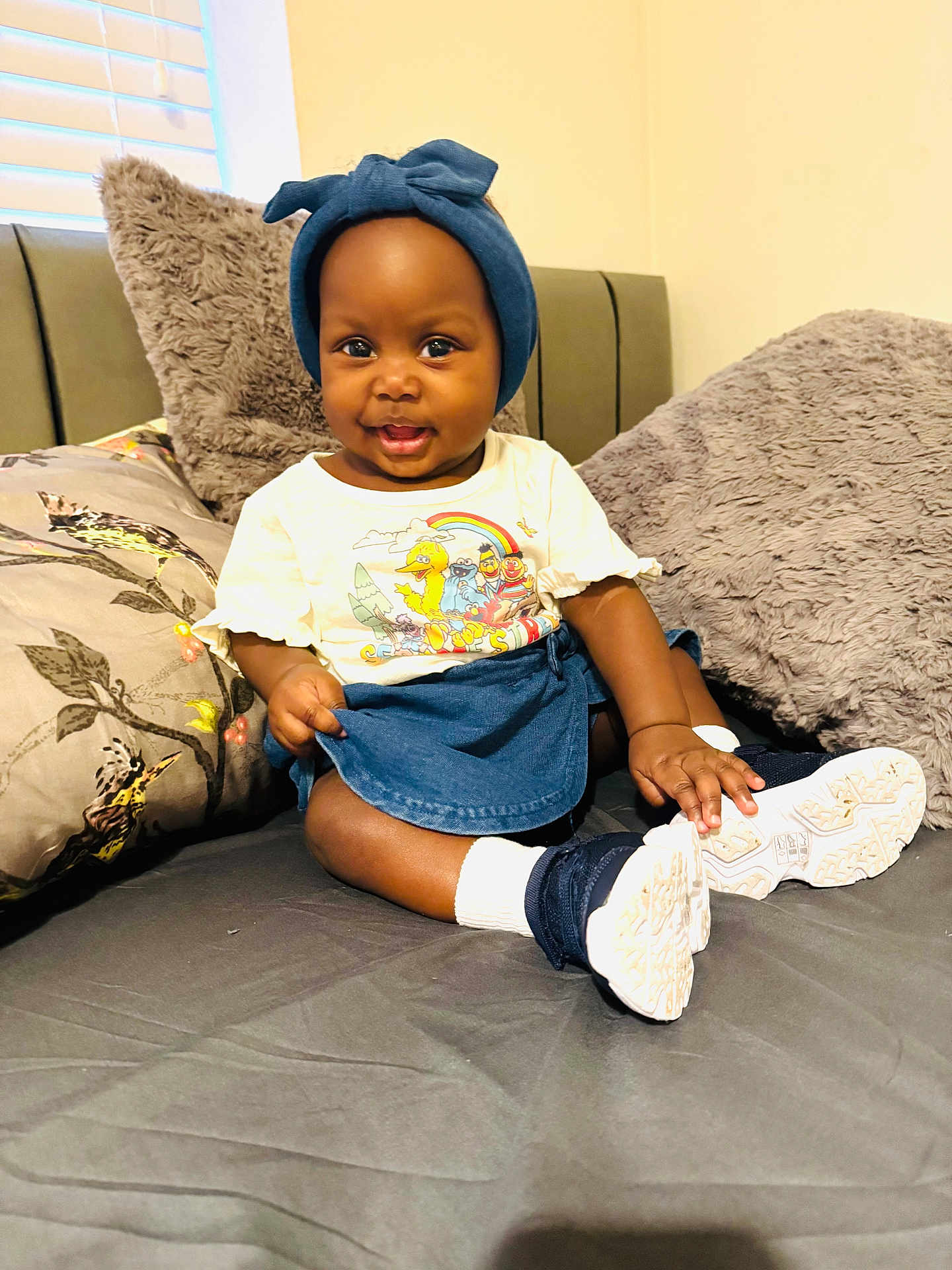 Dinorah is registered to the contest to win money with this photo: baby, child, smiling, bed, pillow, head_wrap, tshirt, shoes, indoor, cute, happy, person, sitting, colorful, clothing, footwear, infant, portrait, cozy, furniture