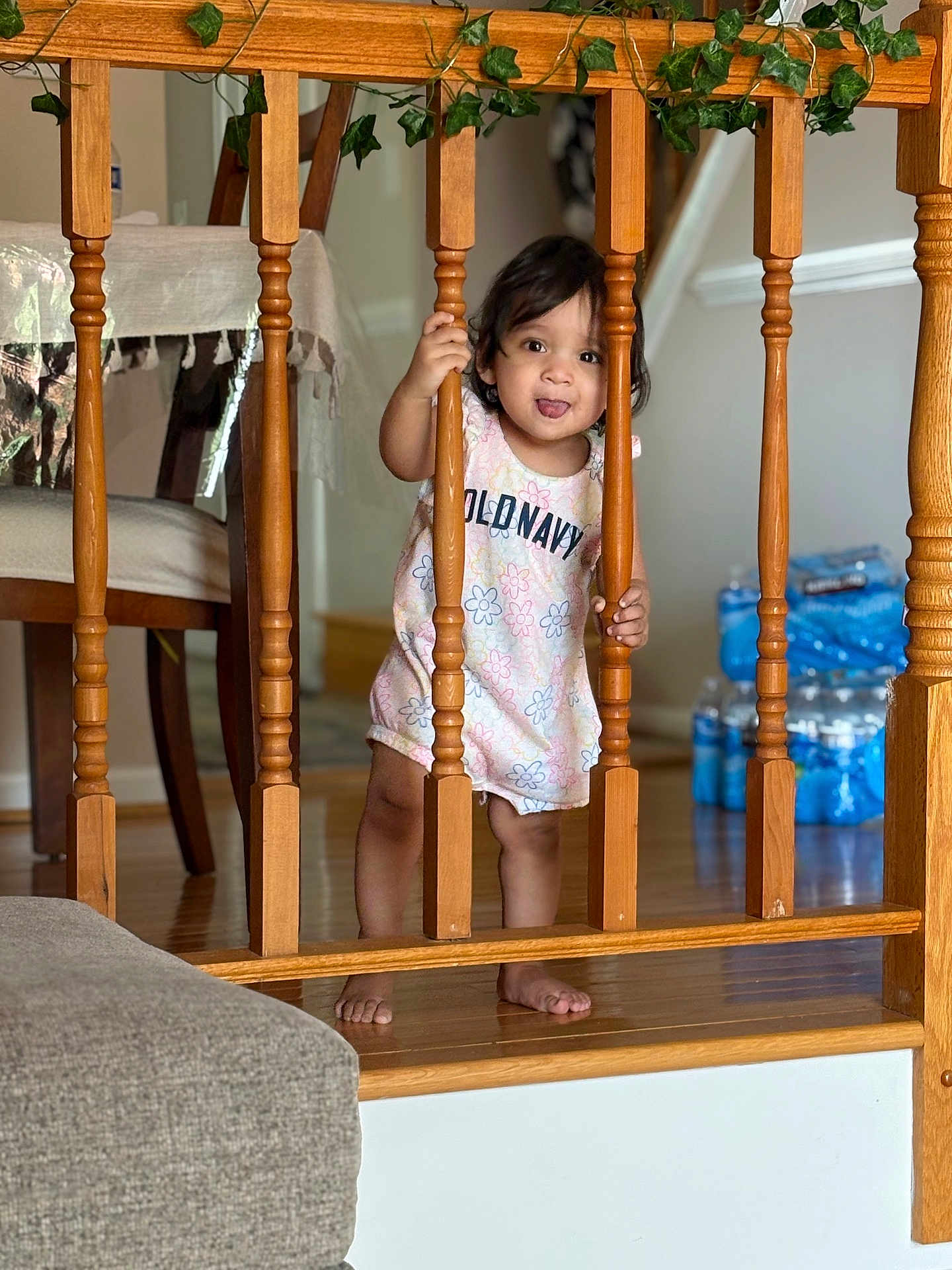Fatima joined the competition — help win amazing prizes! toddler, child, wooden_railings, indoors, flooring, barefoot, dress, expression, tongue_out, chair, tablecloth, water_bottles, wood, hand_holding, smiling, person, home, curious, baby, standing