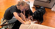 Koda is registered to the contest to win money with this photo: children, dog, reading, book, floor, wooden_floor, couch, indoors, happy, smiling, playful, family, black_dog, casual_clothing, home, interaction, fun, group, child, pet