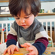 Tom participe au concours pour gagner de l'argent avec cette photo : toddler, child, toy, indoor, sweater, hands, face, playing, educational_toy, table, furniture, room, shelf, picture_frame, curly_hair, concentration, colorful, baby, cute, learning