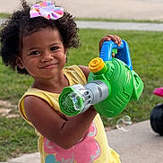 Lainey is registered to the contest to win money with this photo: child, smiling, curly_hair, bow, yellow_shirt, flower_print, bubble_blower, bubbles, outdoor, grass, pavement, playful, toy, summer_clothing, happy, person, cute, colorful, young, casual
