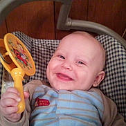 Zach is registered to the contest to win money with this photo: baby, smile, toy, rattle, infant, happy, clothing, blue, striped, hand, face, seat, checkered, wooden_wall, indoor, child, cute, person, young, cozy