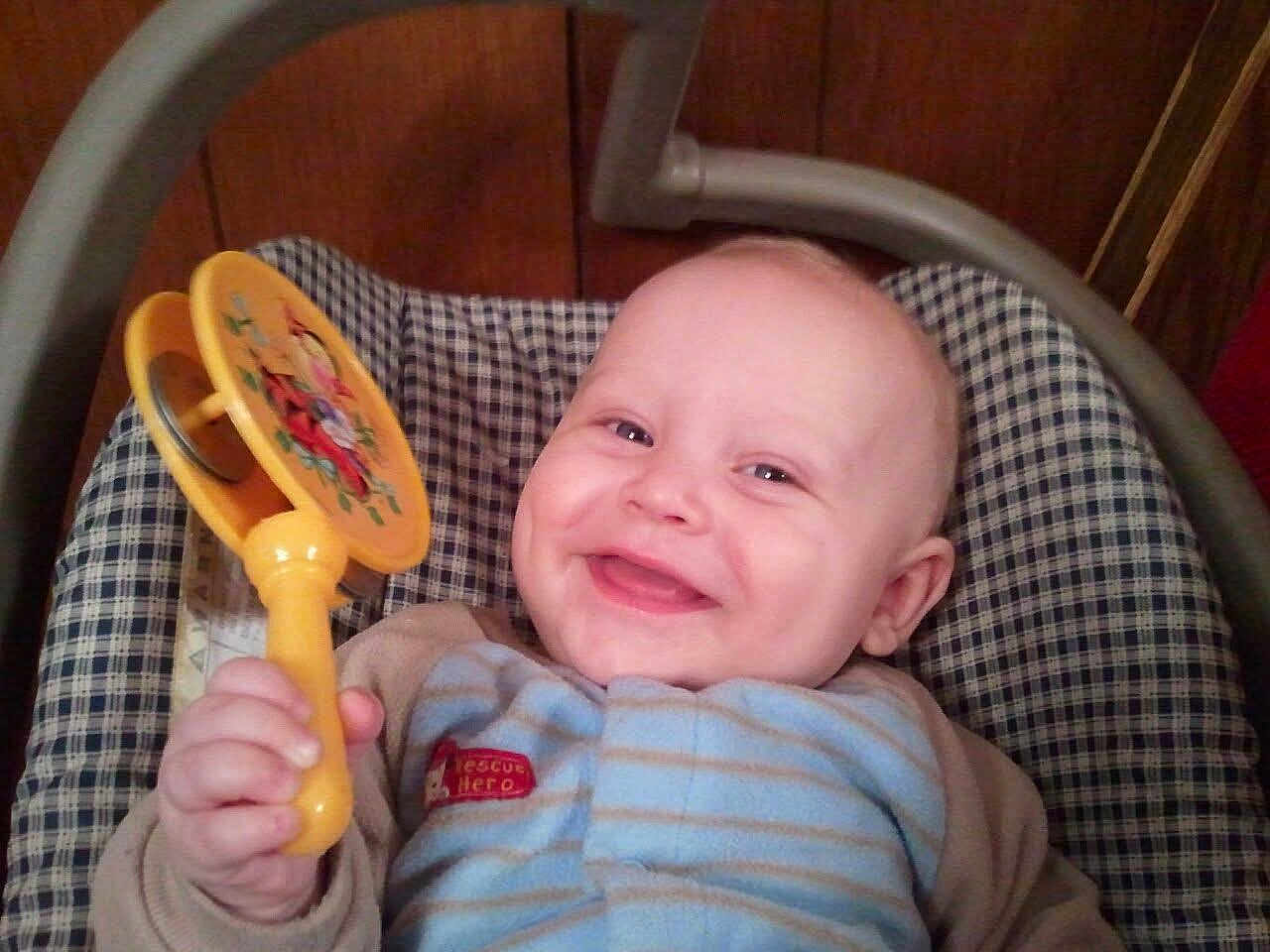 Zach is registered to the contest to win money with this photo: baby, smile, toy, rattle, infant, happy, clothing, blue, striped, hand, face, seat, checkered, wooden_wall, indoor, child, cute, person, young, cozy