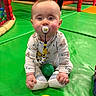 baby, pacifier, green_floor, yellow_ball, green_ball, play_area, child, indoor, crawling, clothing, toy, person, face, infant, colorful, cute, playground, sitting, smiling, footwear