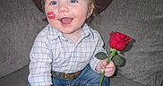 Skyler is registered to the contest to win money with this photo: blue_eyes, boots, casual_clothing, child, couch, cowboy_hat, cute, happy, holding_object, home, indoor, lipstick_mark, person, plaid_shirt, portrait, rose, sitting, smiling, toddler, young_child