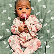 Nina is registered to the contest to win money with this photo: baby, child, comfort, cute, expression, face, floral_pattern, footed_pajamas, green_blanket, hand, indoors, infant, leaf_pattern, onesie, pacifier, pink_clothing, portrait, sitting, soft_texture, young_child