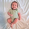 baby, infant, child, smiling, green_onesie, striped_clothing, dinosaur_print, lying_down, white_sheet, happy, cute, portrait, skin, face, head, hands, legs, feet, indoors, soft_lighting