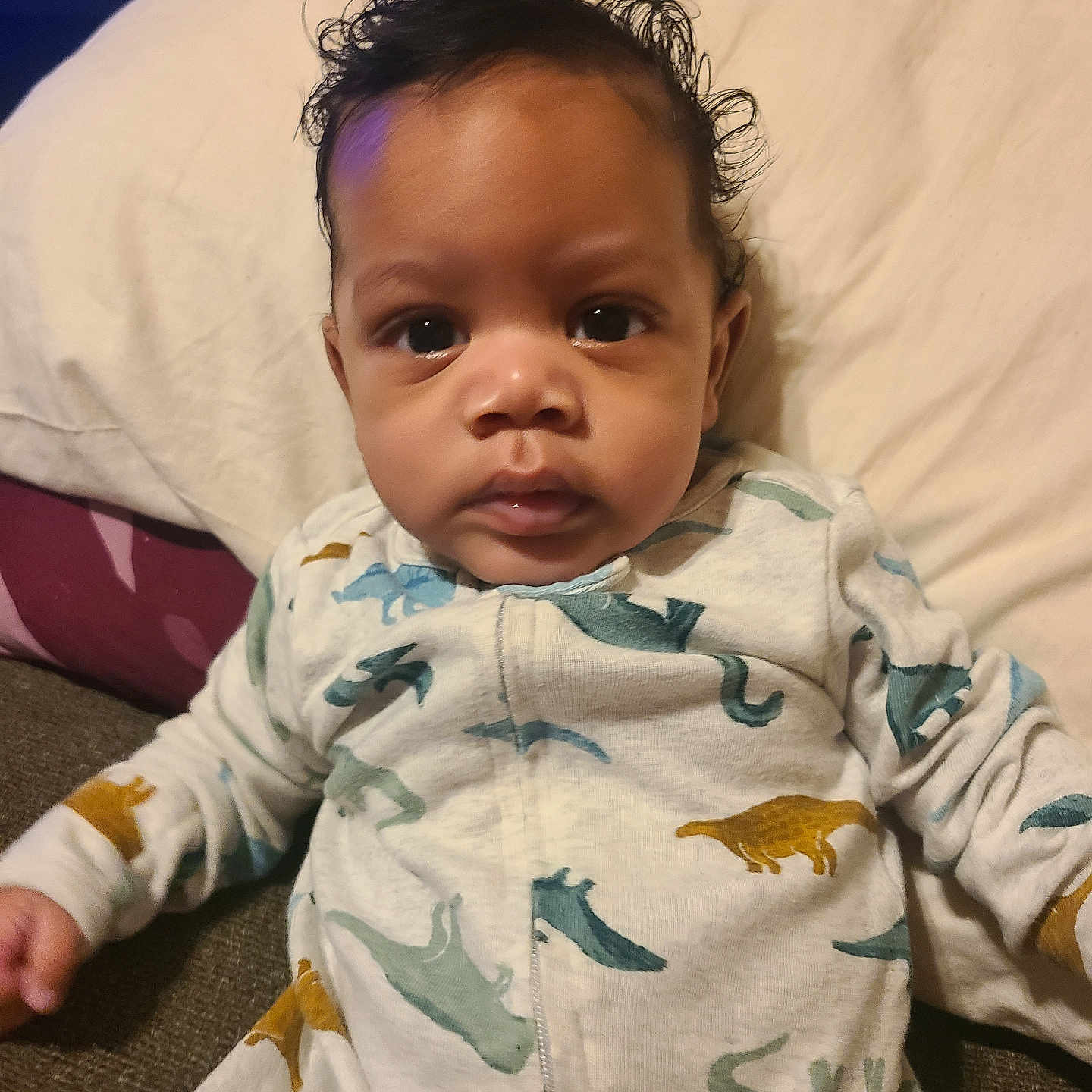 Otao is registered to the contest to win money with this photo: baby, big_eyes, blanket, cheeks, child, close_up, couch, cozy, curly_hair, dinosaur_pattern, face, hands, indoors, infant, onesie, pillow, portrait, romper, sitting, skin