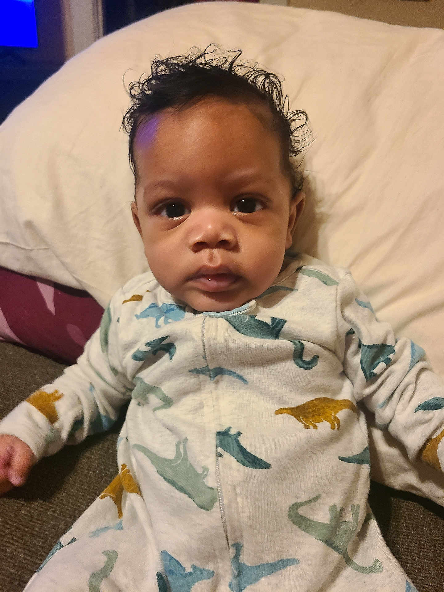 Otao is registered to the contest to win money with this photo: baby, infant, child, onesie, dinosaur_pattern, curly_hair, big_eyes, portrait, close_up, pillow, couch, blanket, romper, sitting, indoors, face, cheeks, hands, skin, cozy