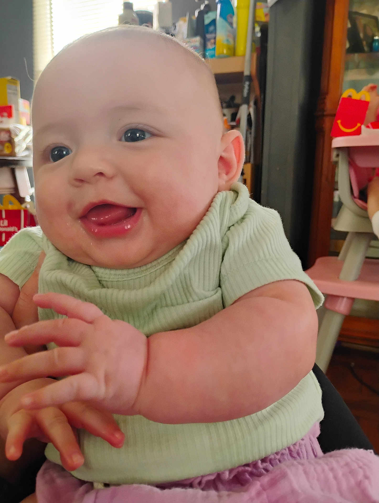 Ella is registered to the contest to win money with this photo: baby, child, smiling, happy, indoor, person, clothing, green_shirt, pink_pants, face, hands, cheeks, bright_eyes, furniture, high_chair, happy_meal_box, household_items, background, closeup, portrait