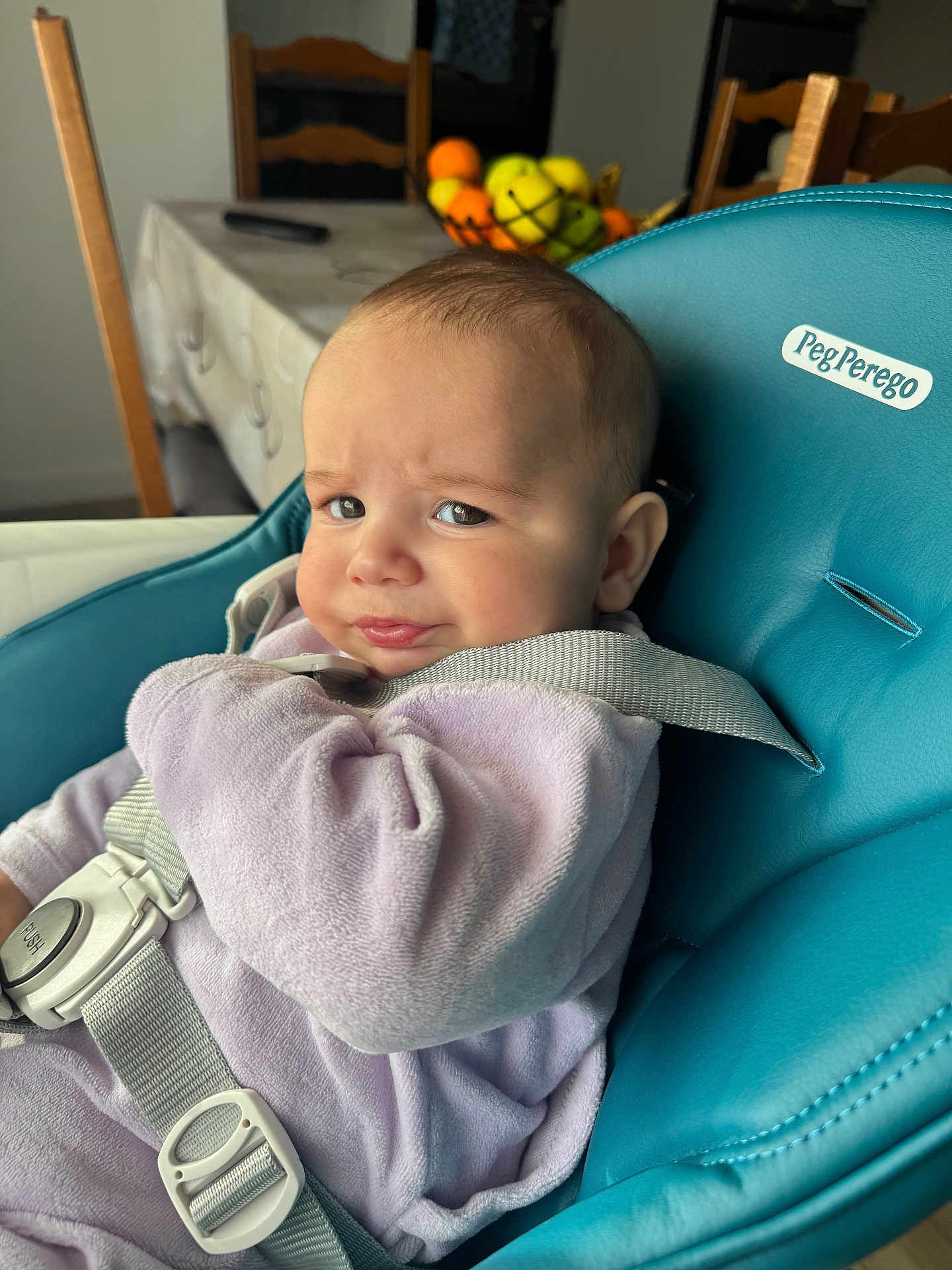 Ionuț Elisei a rejoint le concours — aidez-le/la à gagner de superbes lots ! baby, child, high_chair, purple_clothing, teal_chair, face, expression, indoor, dining_room, fruit_basket, wooden_chairs, table, straps, onesie, person, infant, seat, curious, candid, home