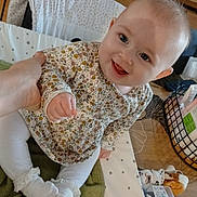 Joy a rejoint le concours — aidez-le/la à gagner de superbes lots ! baby, infant, smiling, sitting, floral_dress, white_socks, green_towel, crib_bumper, parent_hand, wooden_drawer, baby_toy, storage_basket, cotton_swabs_box, indoor, close_up, bright_eyes, happy_expression, cute, table_surface, patterned_sheet