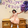 child, girl, birthday, balloon, number_three, pink_dress, cabinet, wooden_furniture, party_decoration, banner, butterflies, celebration, socks, shoes, indoor, smiling, curly_hair, necklace, wall, festive