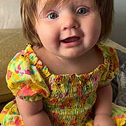 Desiona is registered to the contest to win money with this photo: baby, child, blue_eyes, light_brown_hair, yellow_dress, floral_pattern, cartoon_print, sitting, bed, blanket, curious, smiling, chubby_cheeks, infant, cute, indoors, portrait, person, soft_lighting, young_child