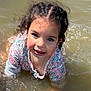 child, water, smiling, playing, outdoor, curly_hair, floral_shirt, face, happy, sunlight, nature, young_child, portrait, joy, summer, water_splash, casual_clothing, necklace, daylight, fun