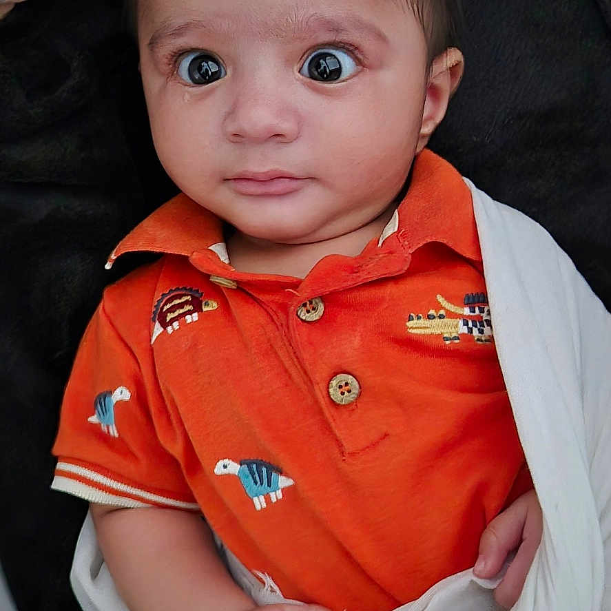 Kartik joined the competition — help win amazing prizes! baby, blanket, child, curious, cute, dinosaur_print, face, hand, head, indoors, infant, orange_shirt, person, portrait, skin, soft_texture, white_cloth, wide_eyes, wrapped, young