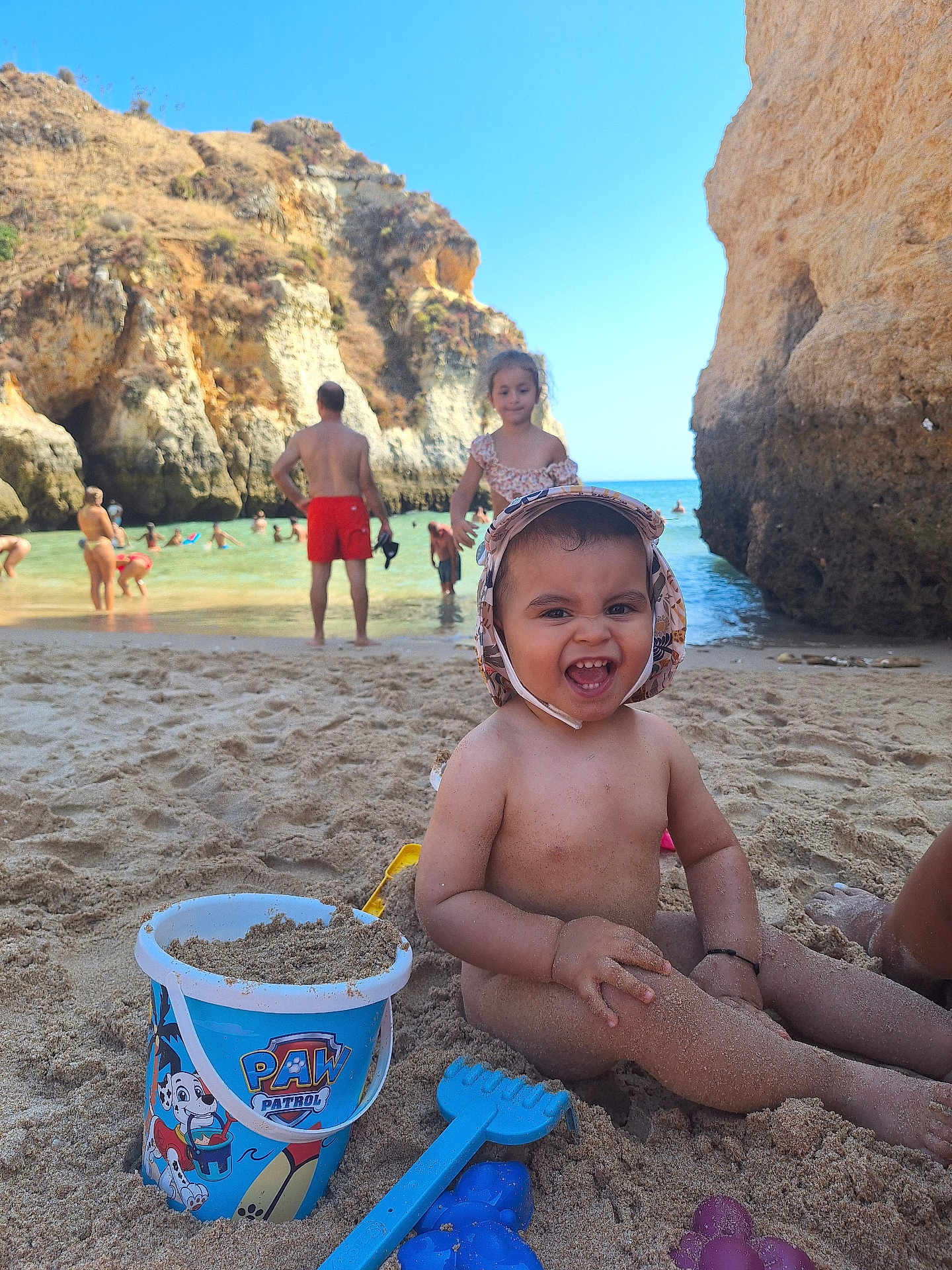 Abel participe au concours pour gagner de l'argent avec cette photo : toddler, child, beach, sand, bucket, toy, smiling, sunny, water, rocks, cliffs, people, swimming, summer, vacation, play, happy, family, outdoor, hat
