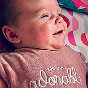 Maddy participe au concours pour gagner de l'argent avec cette photo : adorable, baby, child, close_up, clothing, cute, ear, face, happy, head, indoor, infant, patterned_background, person, pink_sweater, portrait, skin, smiling, text_on_clothing, young