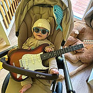 Aaron a rejoint le concours — aidez-le/la à gagner de superbes lots ! baby, stroller, guitar, sunglasses, hat, plush_toy, floor, wood_floor, indoor, child, person, furniture, window, light, toy, blanket, foot, hand, clothing, seat