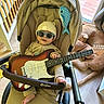 baby, stroller, guitar, sunglasses, hat, plush_toy, floor, wood_floor, indoor, child, person, furniture, window, light, toy, blanket, foot, hand, clothing, seat
