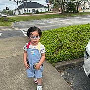 Dwayne is registered to the contest to win money with this photo: toddler, child, glasses, overalls, sidewalk, green_bush, car, suburban, street, tree, grass, cloudy_sky, footwear, person, outdoor, walking, building, residential, casual_clothing, daytime