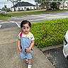 toddler, child, glasses, overalls, sidewalk, green_bush, car, suburban, street, tree, grass, cloudy_sky, footwear, person, outdoor, walking, building, residential, casual_clothing, daytime