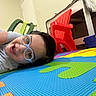 Dwayne is registered to the contest to win money with this photo: bright_colors, casual_clothing, child, colorful_mat, face, floor, foam_puzzle, glasses, green_chair, happy, head, home_interior, indoor, lying_down, person, playroom, red_chair, smiling, toy_storage, young_child