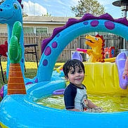 Dwayne is registered to the contest to win money with this photo: backyard, blue, casual_clothing, child, dinosaur, fence, fun, grass, green, inflatable_pool, outdoor, play, purple, smile, summer, sunny, toy, water, wet_hair, yellow