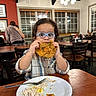 blinds, casual, chair, child, dining, eating, food, fork, fried_chicken, glasses, indoor, lighting, meal, person, plaid_shirt, plate, restaurant, table, window, wooden_furniture