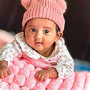 Eva is registered to the contest to win money with this photo: baby, infant, pink_hat, pom_poms, blanket, knitted, floral_clothing, cute, portrait, child, wide_eyes, soft_texture, indoor, resting, adorable, young_child, close_up, warm_colors, cozy, soft_focus