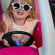 Gracelyn is registered to the contest to win money with this photo: child, girl, sunglasses, toy_car, pink, smile, blonde_hair, steering_wheel, indoor, play, happy, cute, person, fashion, accessory, casual, fun, portrait, seat, hands