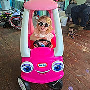 Gracelyn is registered to the contest to win money with this photo: adult, bicycle, bucket, casual_clothing, child, clutter, floor, indoor, person, pink, plastic, playroom, seat, smiling, steering_wheel, sunglasses, toy, toy_car, wall_art, wooden_floor