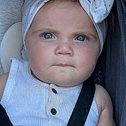 Evie is registered to the contest to win money with this photo: baby, child, blue_eyes, headband, bow, white_clothing, car_seat, straps, serious_expression, close_up, infant, face, portrait, sitting, cute, skin, hair, indoors, seatbelt, young_child