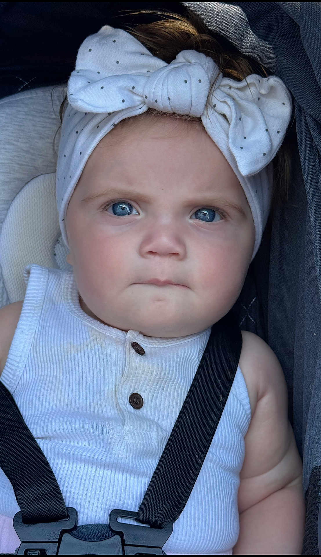 Evie is registered to the contest to win money with this photo: baby, child, blue_eyes, headband, bow, white_clothing, car_seat, straps, serious_expression, close_up, infant, face, portrait, sitting, cute, skin, hair, indoors, seatbelt, young_child