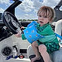 child, toddler, boat, steering_wheel, floaties, wet_hair, green_swimsuit, blue_floaties, seat, sky, clouds, sunny, barefoot, water_safety, outdoor, summer, playful, curious, person, vehicle
