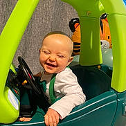 Anakin is registered to the contest to win money with this photo: baby, child, toy_car, green, teal, steering_wheel, laughing, happy, indoor, balloon, tiger_balloon, clothing, white_shirt, overalls, hand, face, head, play, smile, person