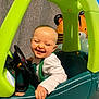 baby, child, toy_car, green, teal, steering_wheel, laughing, happy, indoor, balloon, tiger_balloon, clothing, white_shirt, overalls, hand, face, head, play, smile, person