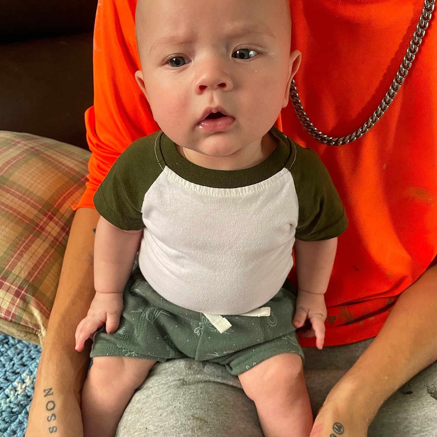 Hudsyn joined the competition — help win amazing prizes! adult, baby, casual_clothing, chain_necklace, child, curious_expression, face, hand, indoor, knitted_blanket, lap, legs, orange_shirt, person, plaid_pillow, shirt, shorts, sitting, skin, tattoo