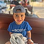 child, boy, cap, backwards_cap, blue_tshirt, blanket, smiling, face, hand, outdoor, daylight, person, seat, vehicle, background, casual_clothing, portrait, happy, young_child, closeup