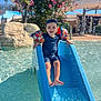 child, boy, water_slide, pool, floaties, blue_slide, water, summer, sunny, cap, swimwear, splash, outdoor, play, happy, nature, greenery, flowers, daylight, recreation