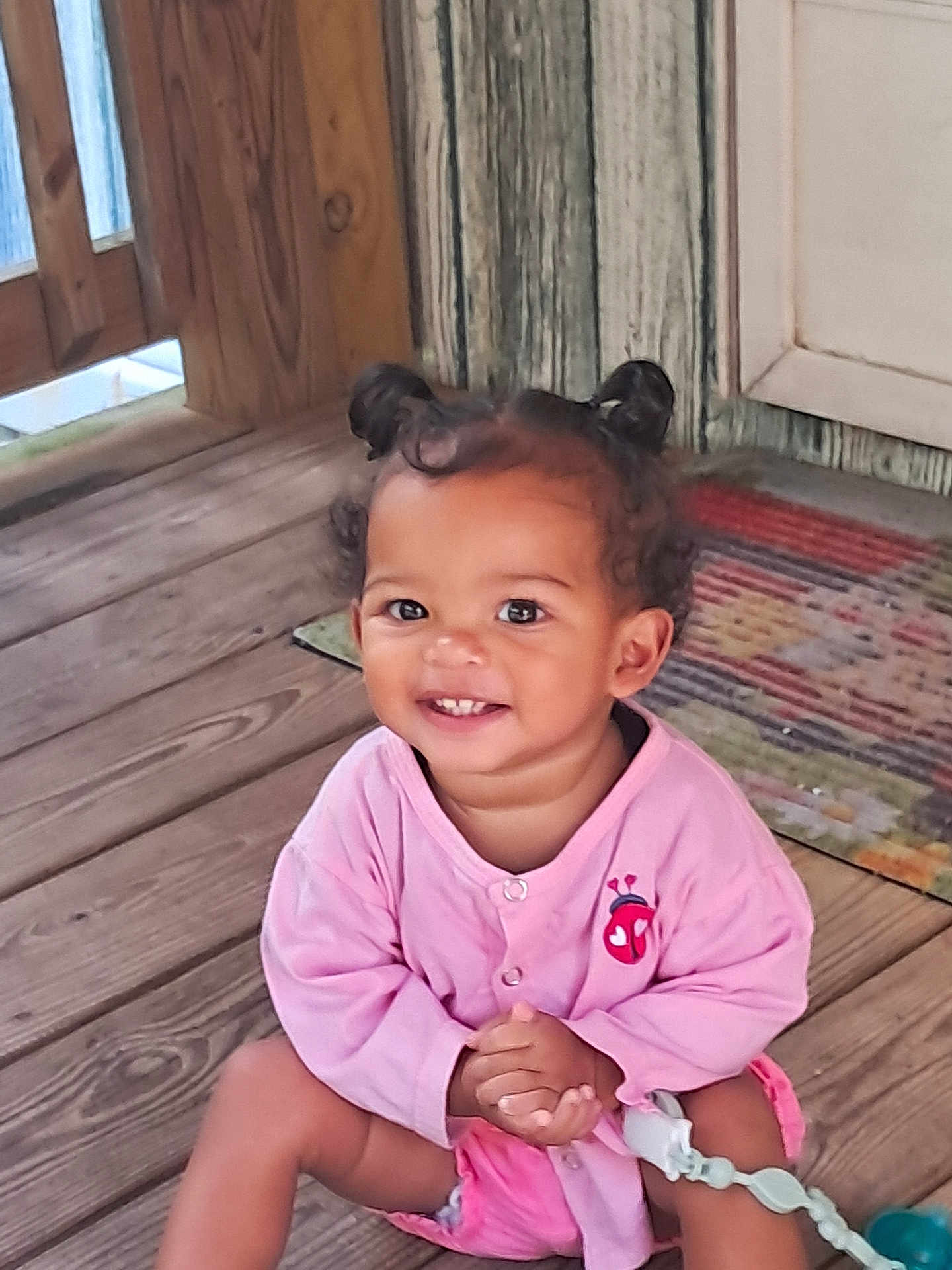 Chianne is registered to the contest to win money with this photo: toddler, child, smiling, pink_clothing, hair_buns, wooden_floor, indoor, cute, happy, baby, person, portrait, sitting, decor, rug, hands_clasped, young_child, casual_clothing, cute_hair, playful