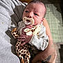 Grayson is registered to the contest to win money with this photo: baby, newborn, infant, pacifier, plush_giraffe, stuffed_animal, holding, hand, arm, tattoo, onesie, blanket, indoor, portrait, gaze, bedside, cozy, wood_furniture, rug, caretaker_arm