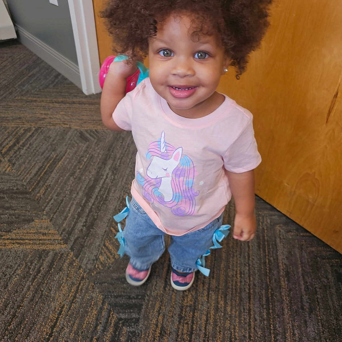 Tori Jones joined the competition — help win amazing prizes! child, toddler, curly_hair, indoor, carpet, pink_shirt, unicorn, jeans, ribbons, smiling, holding_toy, footwear, door, wall, earrings, person, cute, happy, standing, portrait