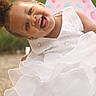 child, toddler, smiling, white_dress, curly_hair, earrings, chair, outdoor, green_background, happy, portrait, cute, joyful, person, fashion, sitting, daylight, young, pretty, fun