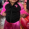 Tori Jones is registered to the contest to win money with this photo: toddler, child, smiling, standing, indoors, wooden_floor, pink_clothing, black_jacket, beads, hair_accessory, sandals, window, furniture, red_inflatable, person, flooring, happy, portrait, room, casual